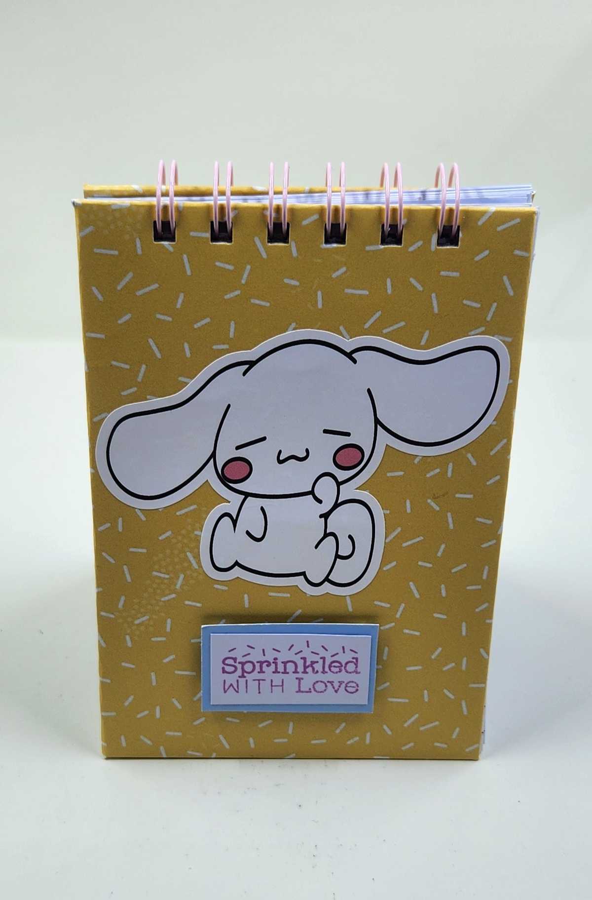 Handmade Cinnamoroll Notepad with yellow sprinkle paper background, Sprinkled with Love sentiment and Cinnamoroll image standing upright.