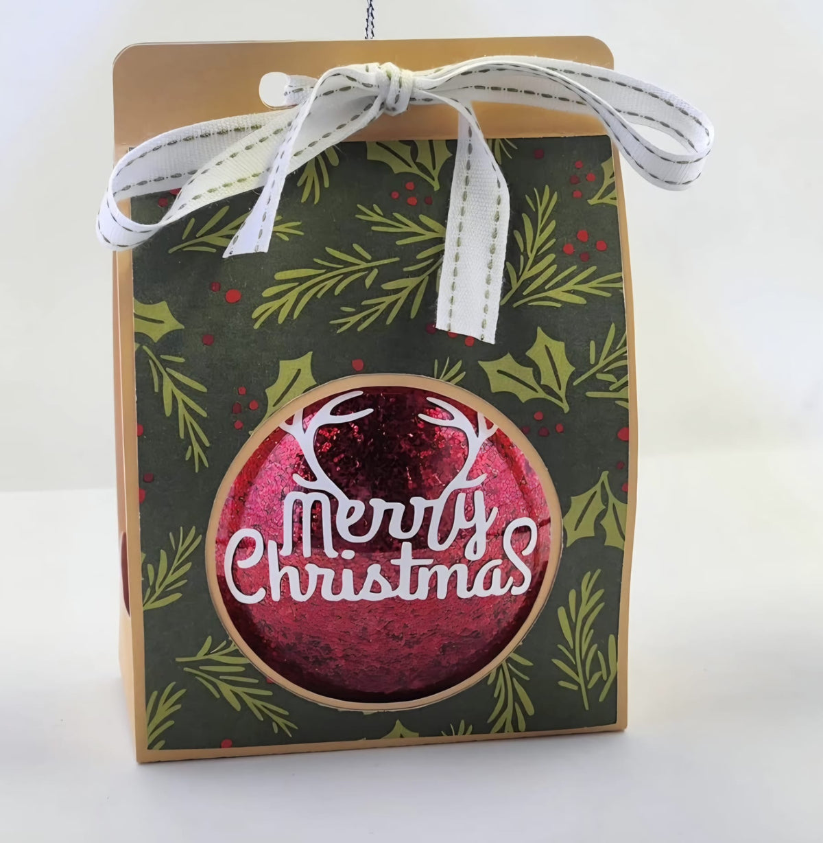 Red glitter Christmas bauble with a white "Merry Christmas" greeting, in a holly-patterned gift box
