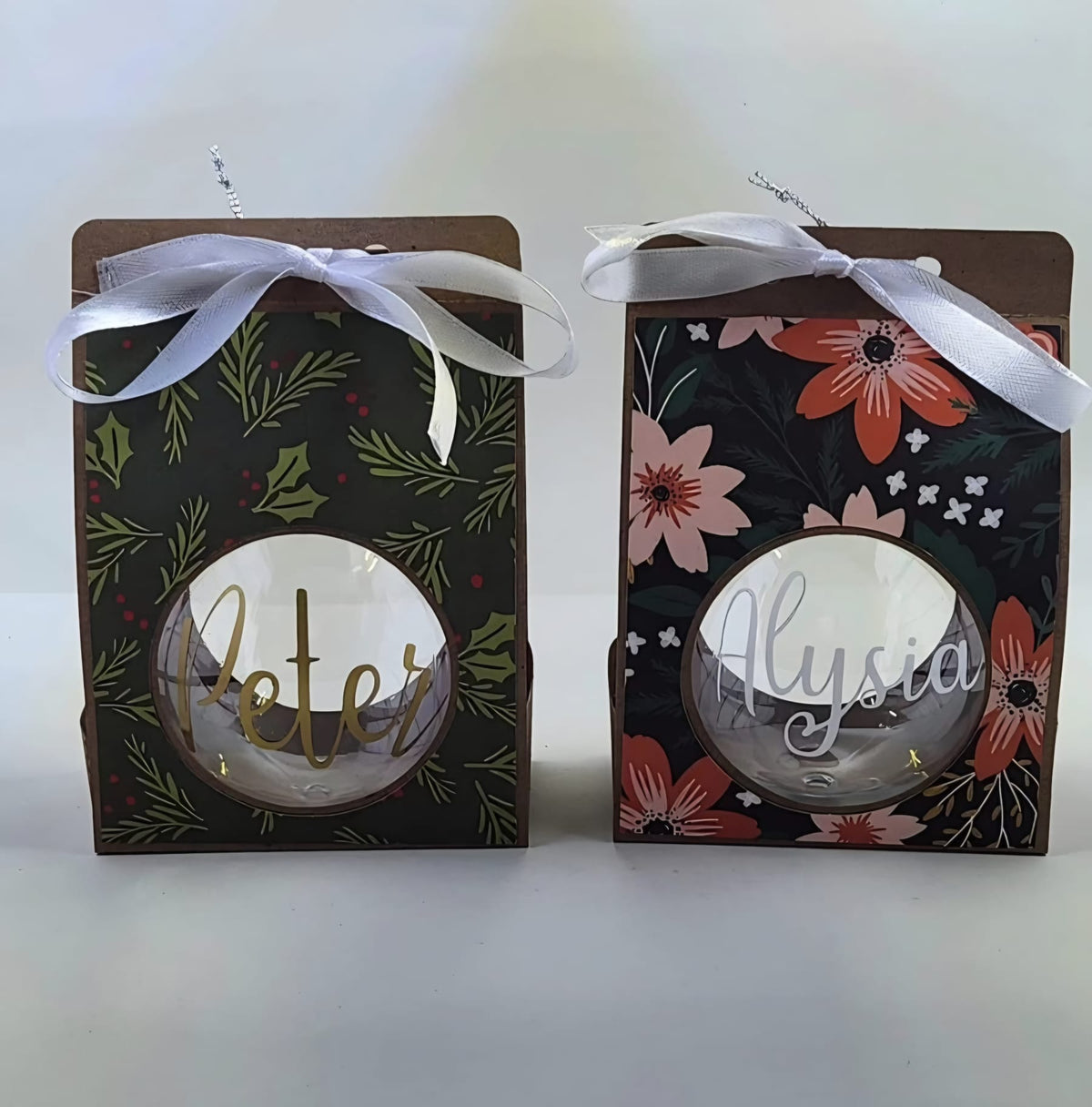 Personalised clear Christmas baubles with the names Peter in gold and Alysia in white, shown in floral and holly gift boxes